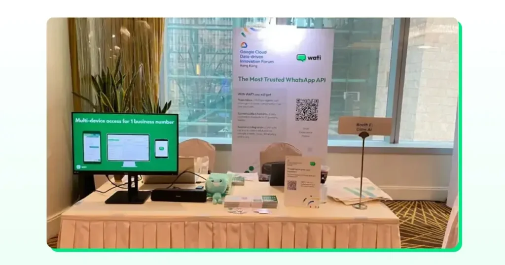 Photo of a Wati event booth displaying a monitor that shows WhatsApp multi-device access for one business number, along with product brochures and branding.